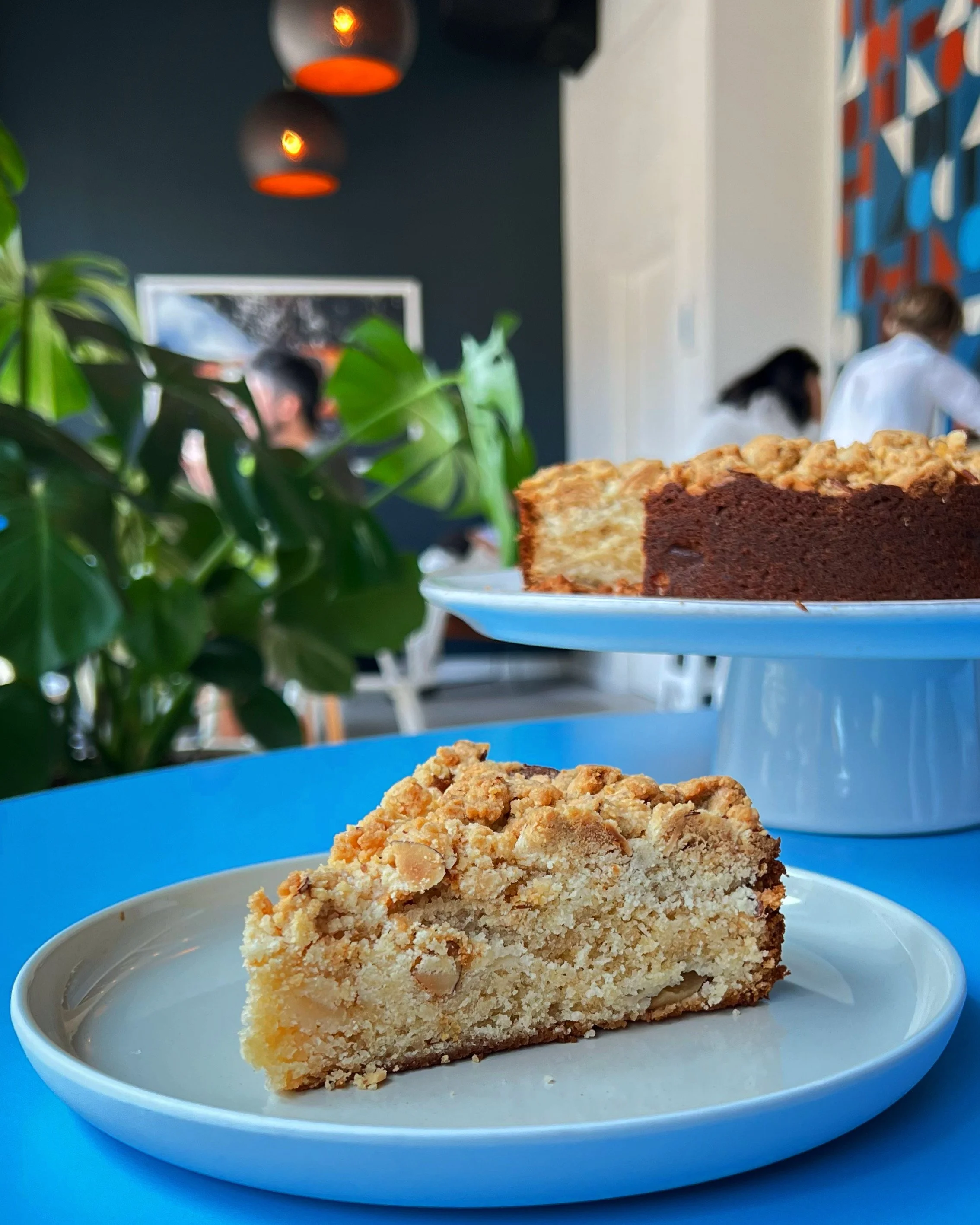 A slice of crumb cake on a small white plate, with two additional slices of cake on a white cake stand in the background, inside a cafe with people and colorful wall art.