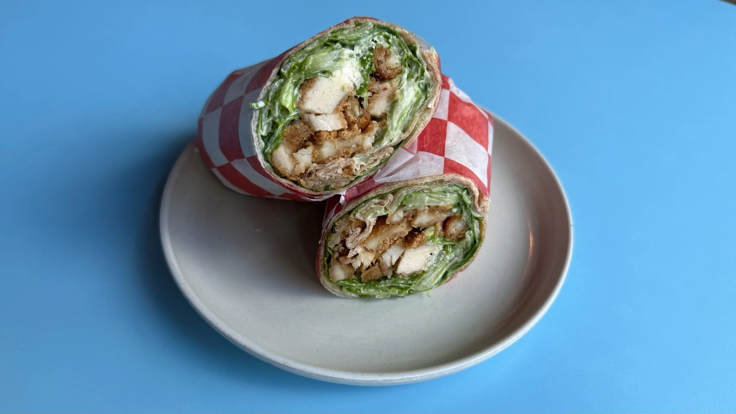 A breakfast burrito cut in half on a white plate, with lettuce, chicken, cheese, and other ingredients inside, wrapped in red and white checkered paper, against a light blue background.