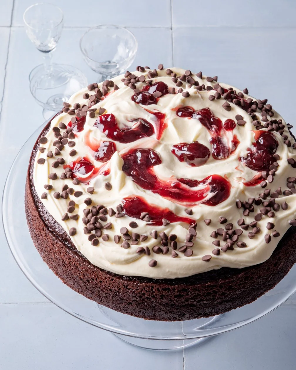Chocolate cake with white frosting, cherry topping, and chocolate chips on top, placed on a glass stand with empty glasses in the background.