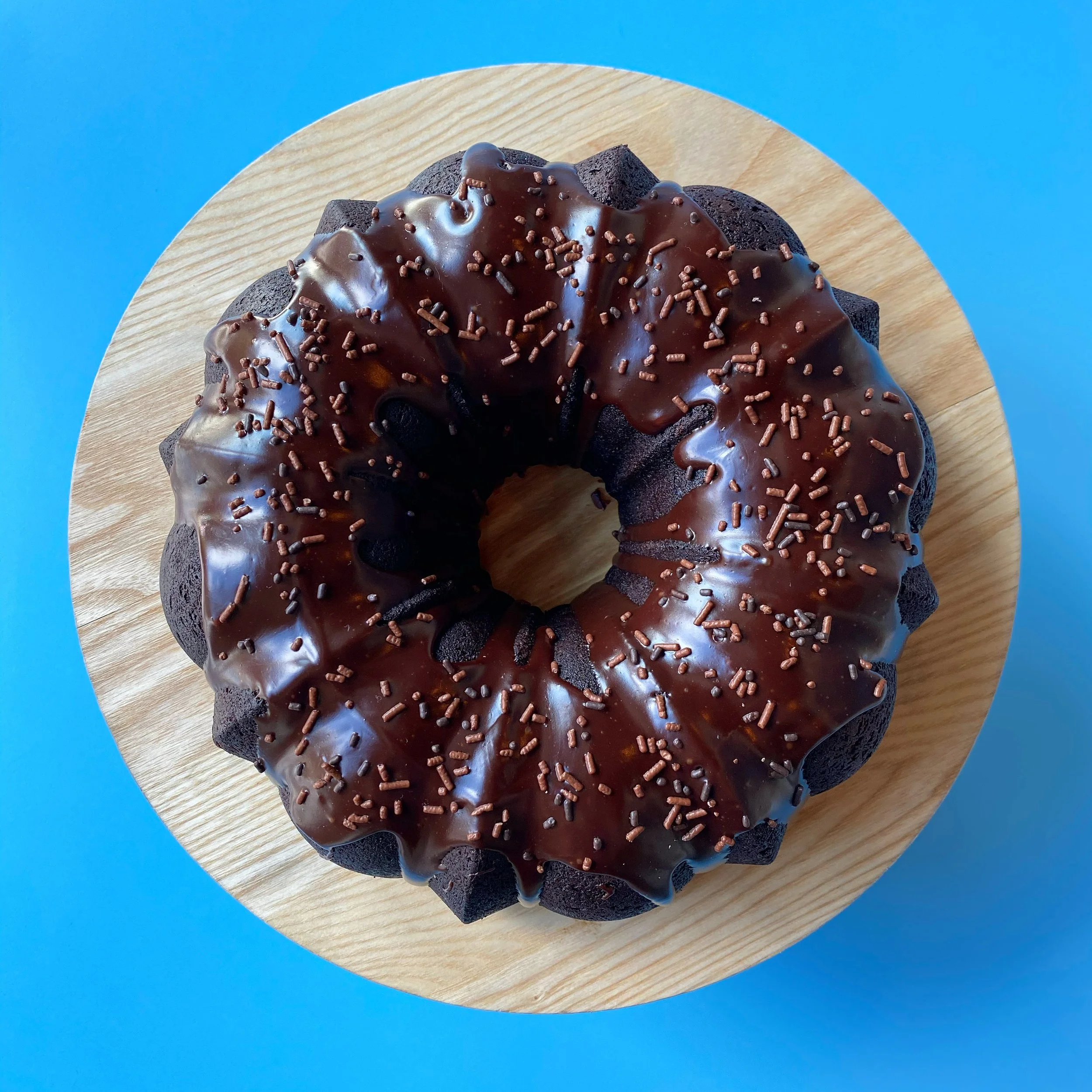 Chocolate glazed bundt cake with sprinkles on a wooden board against a blue background.