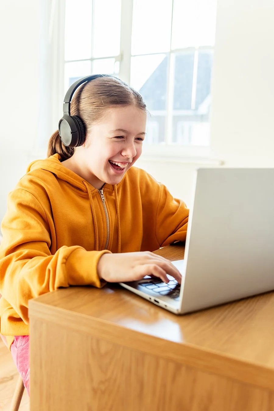 A girl sitting at a wooden table working on a laptop while wearing headphones and a yellow hoodie, using Assistive Technology for improved reading and writing at school after an AT evaluation in Buffalo, NY.