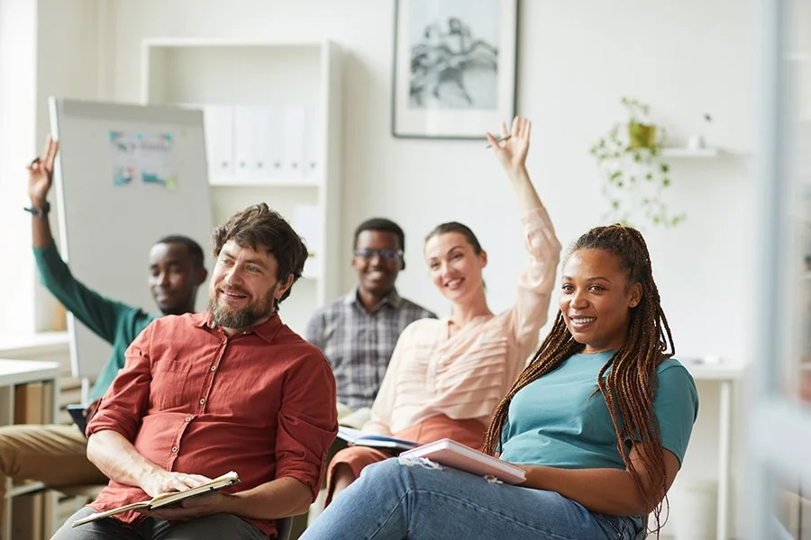 Group of diverse teachers and support staff sitting in a classroom raising hands and smiling during a training for educators on Assistive Technology and AAC in Buffalo, NY.