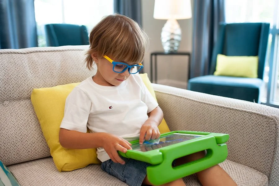 A young boy with glasses sits on a beige couch, using an AAC app on a tablet that he received after an AAC evaluation in Buffalo, NY, in a bright living room with blue chairs and curtains in the background.