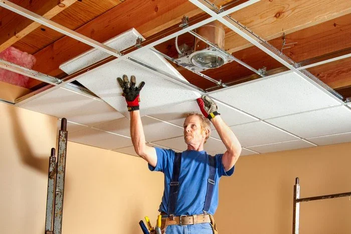 A man installing a dropped ceiling or ceiling tiles in a room with exposed wooden beams on the ceiling. He is standing on the ground, wearing a blue shirt and work gloves.