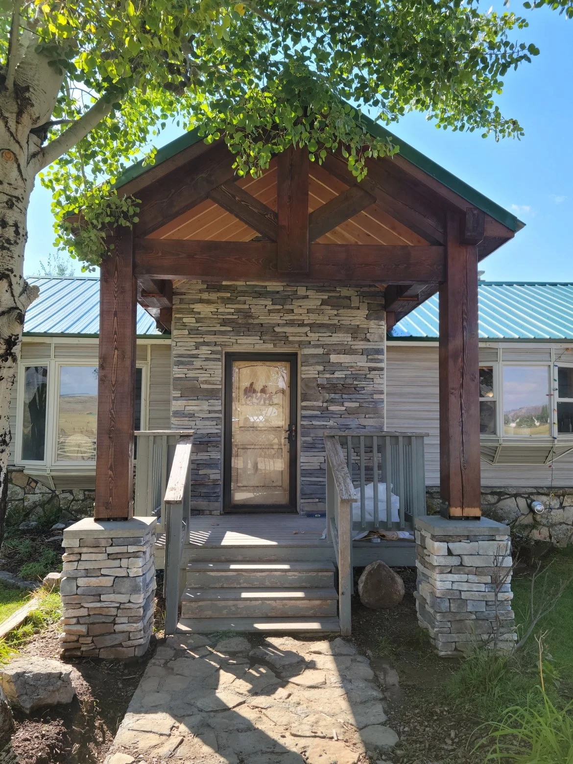 Front view of a house with a small porch and stone steps, a glass door, surrounded by a tree and green grass, with a grassy landscape in the background under a clear blue sky.