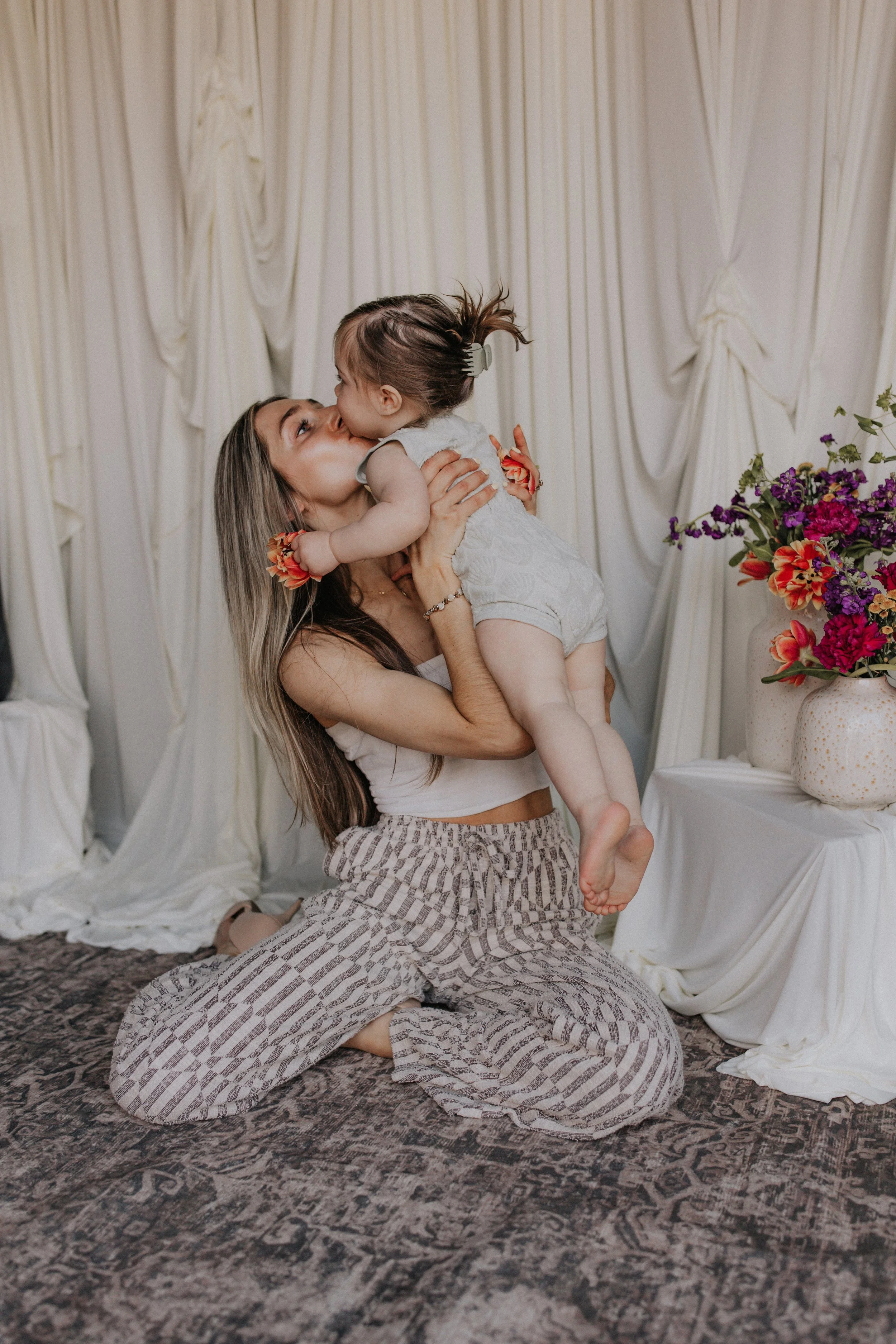 A woman kneeling on a patterned rug, holding a young girl in her arms, both sharing a kiss. The woman is wearing striped pants and a white top, while the girl wears a sleeveless dress. Behind them, there are cream-colored drapes and vases with colorful flowers.