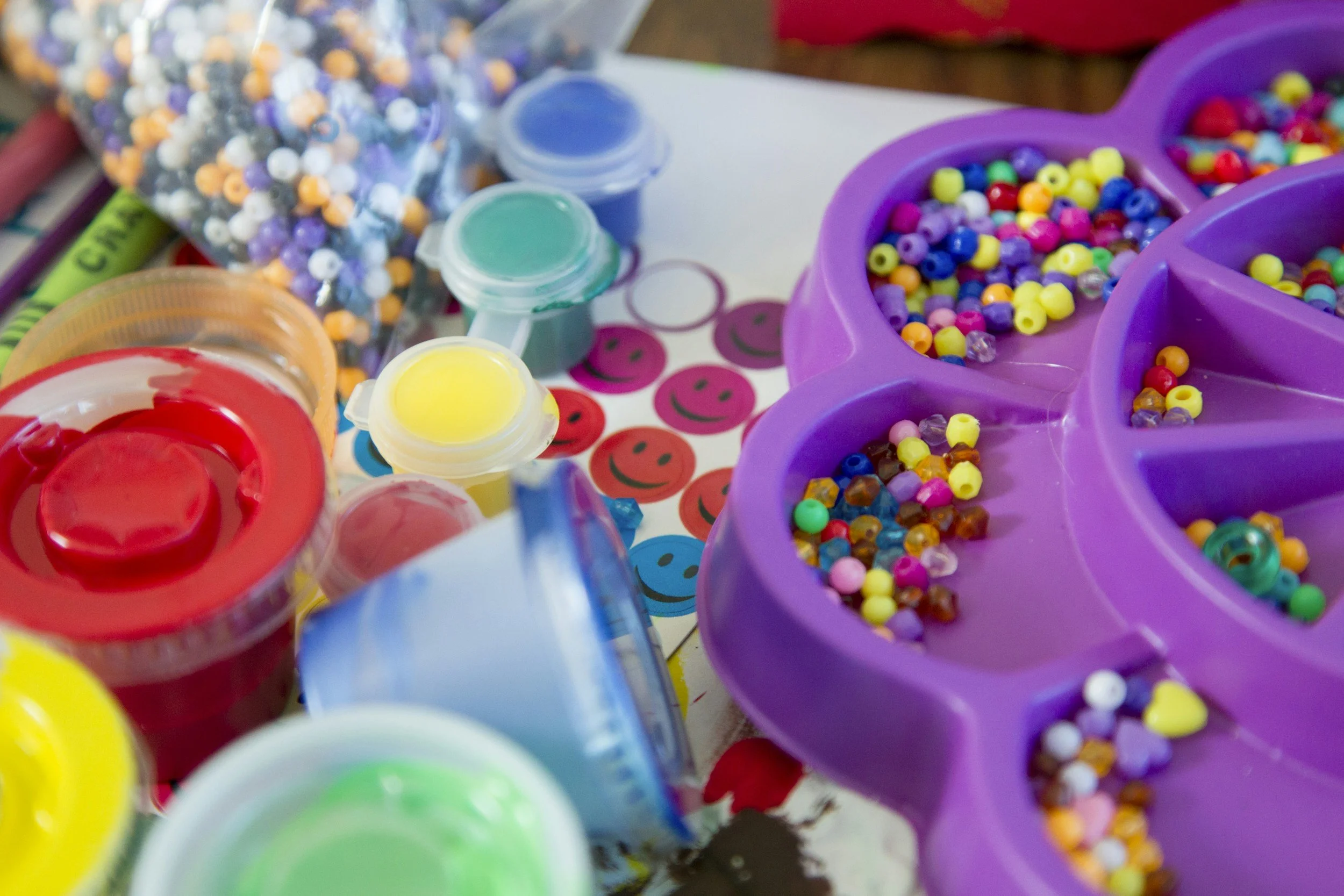 Assorted colorful craft beads, containers of paint, and smiley face stickers arranged on a table, possibly for arts and crafts activity.