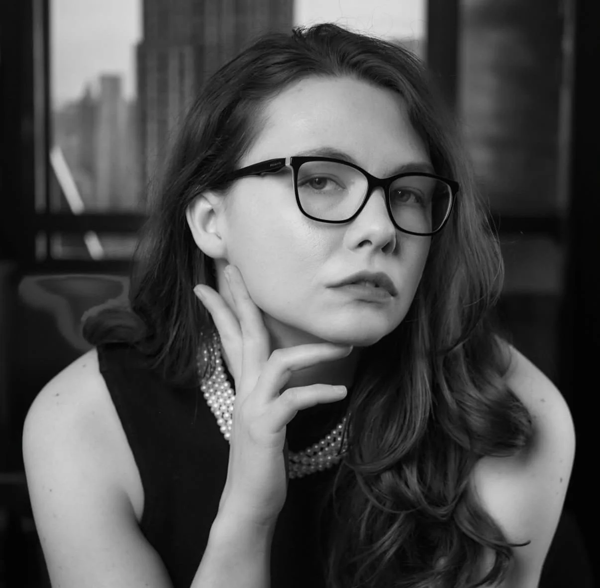 Black and white portrait of a woman with long wavy hair wearing glasses, a pearl necklace, and a sleeveless top, sitting indoors with bookshelves in the background.