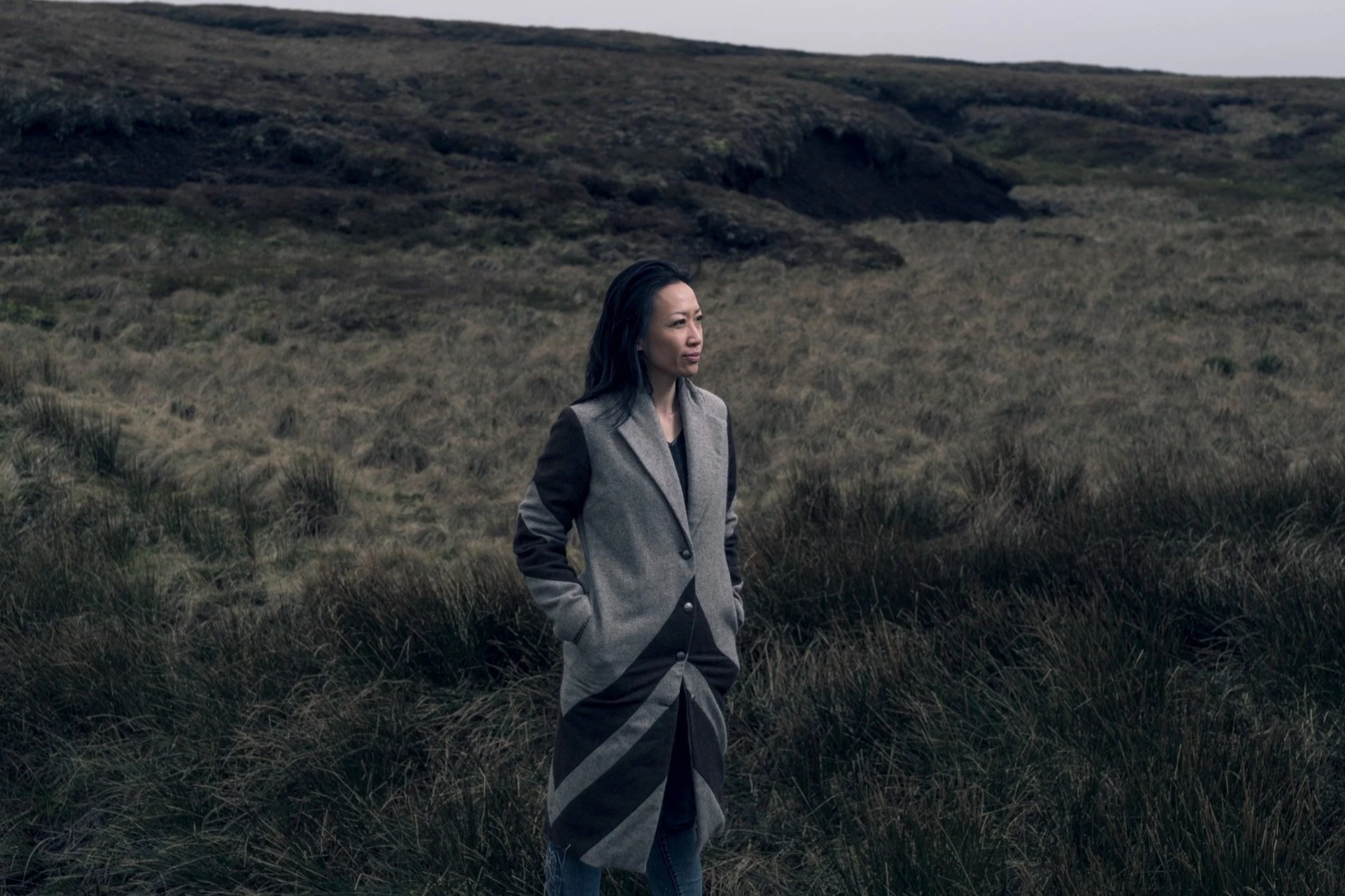 Woman standing in a grassy, hilly landscape with overcast sky, wearing a long, patterned coat and hands in pockets.