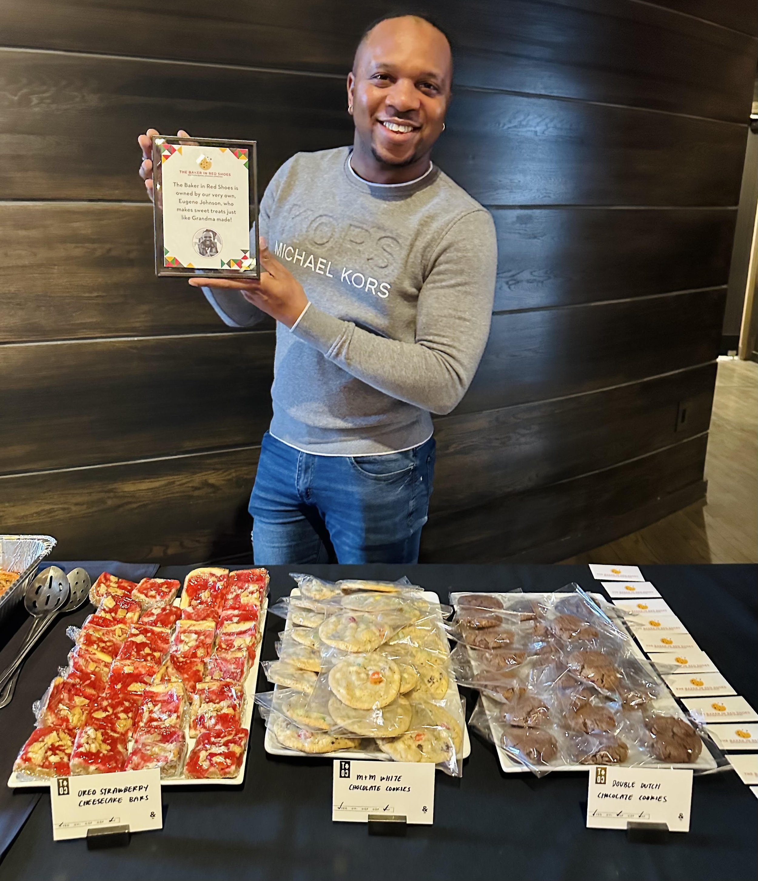 A man smiling and holding a framed certificate or award at a table filled with various cookies, including Oreo strawberry cheesecake bars, white chocolate cookies, and double Dutch chocolate cookies, set against a dark wood-paneled wall.