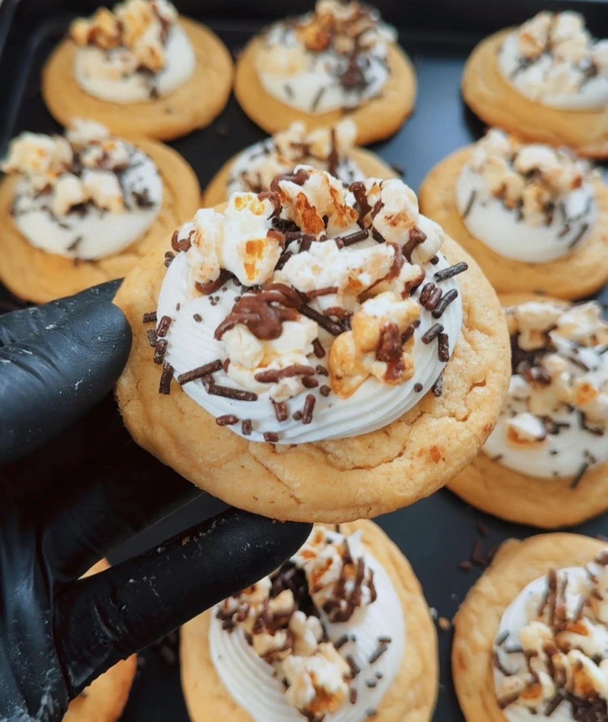 A hand wearing a black glove holding a cookie with white frosting, popcorn, chocolate sprinkles, and drizzled chocolate, with more similar cookies in the background.