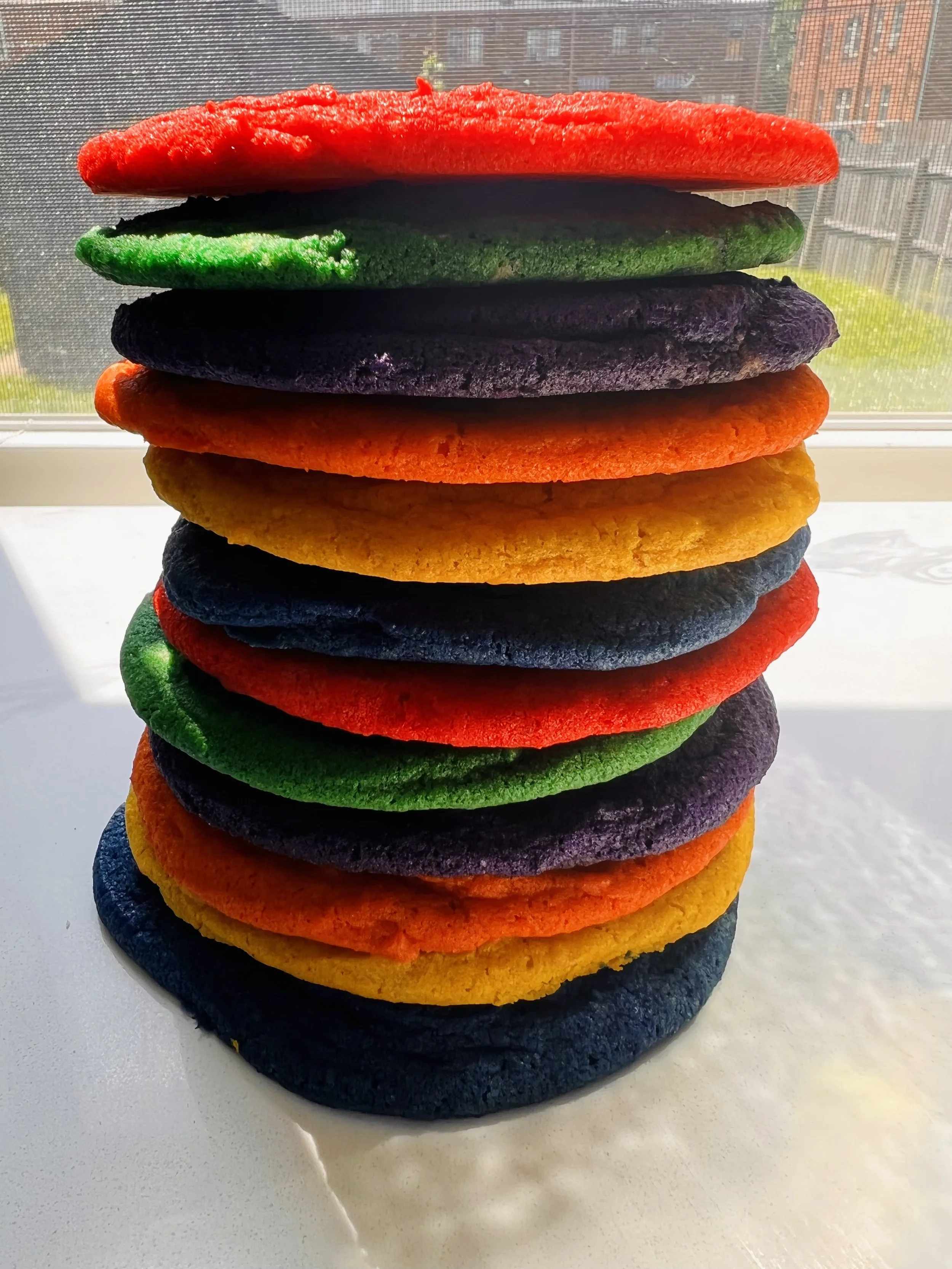 A stack of multi-colored cookies arranged in rainbow order on a white surface near a window with a cityscape background.