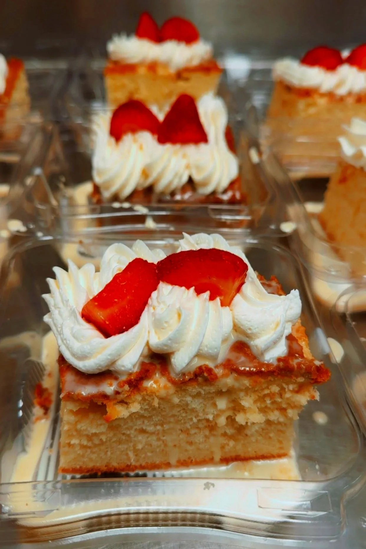 A close-up view of a slice of vanilla cake with whipped cream and fresh strawberry slices on top in a clear plastic container.