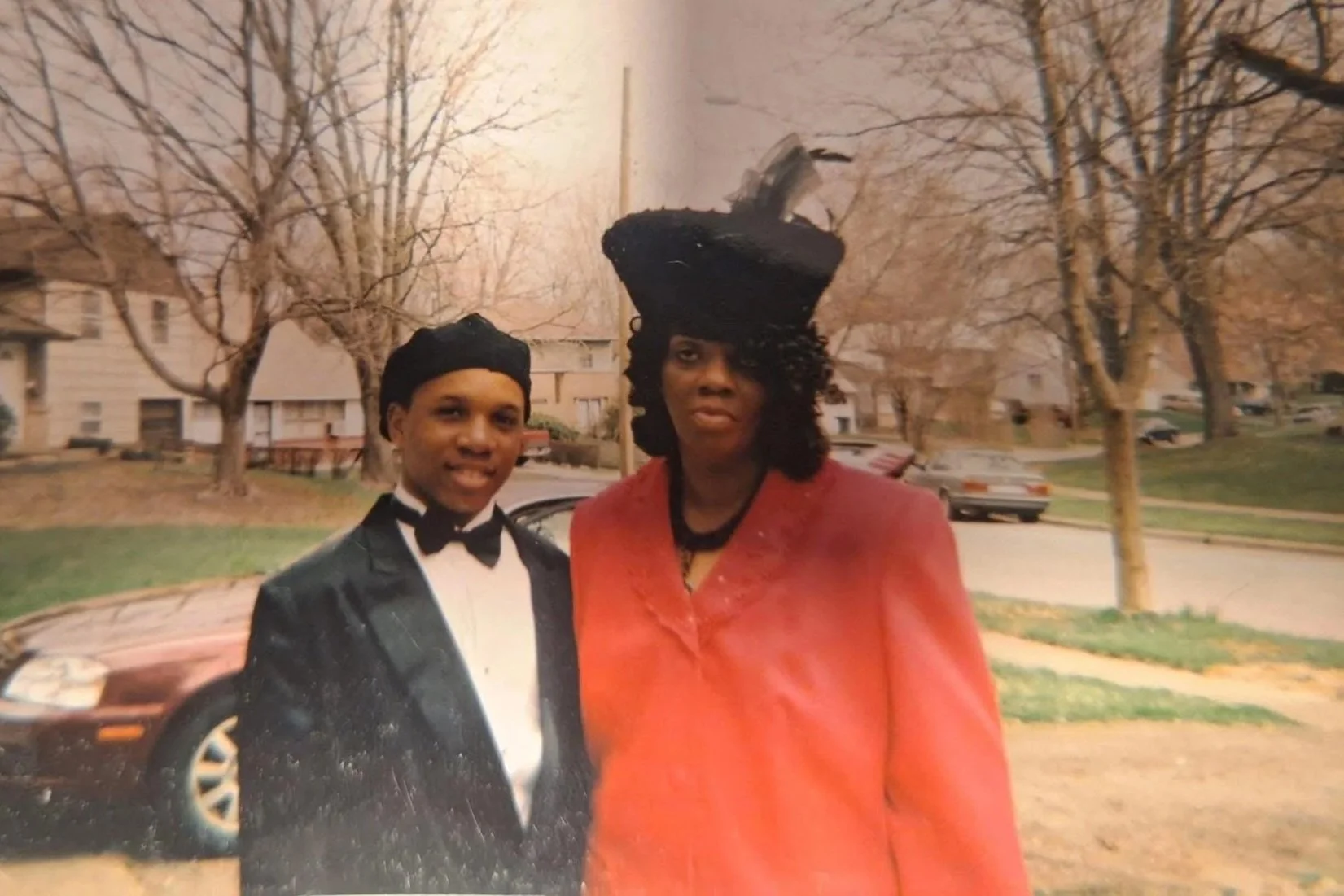 A young man in a tuxedo standing next to an older woman in a bright orange coat and black hat, outdoors in a residential neighborhood with leafless trees and parked cars.
