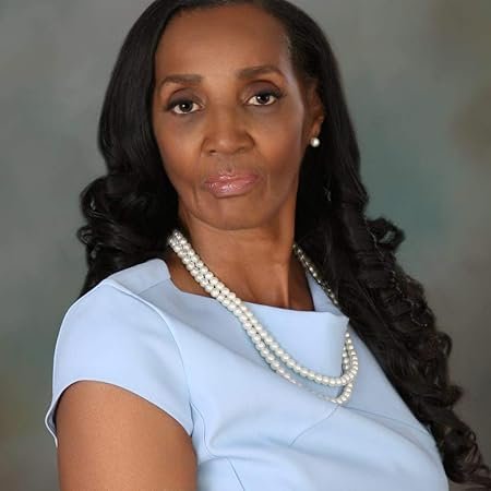 A mature woman with long black curly hair, wearing a light blue top, pearl necklace, and earrings, posing against a neutral background.