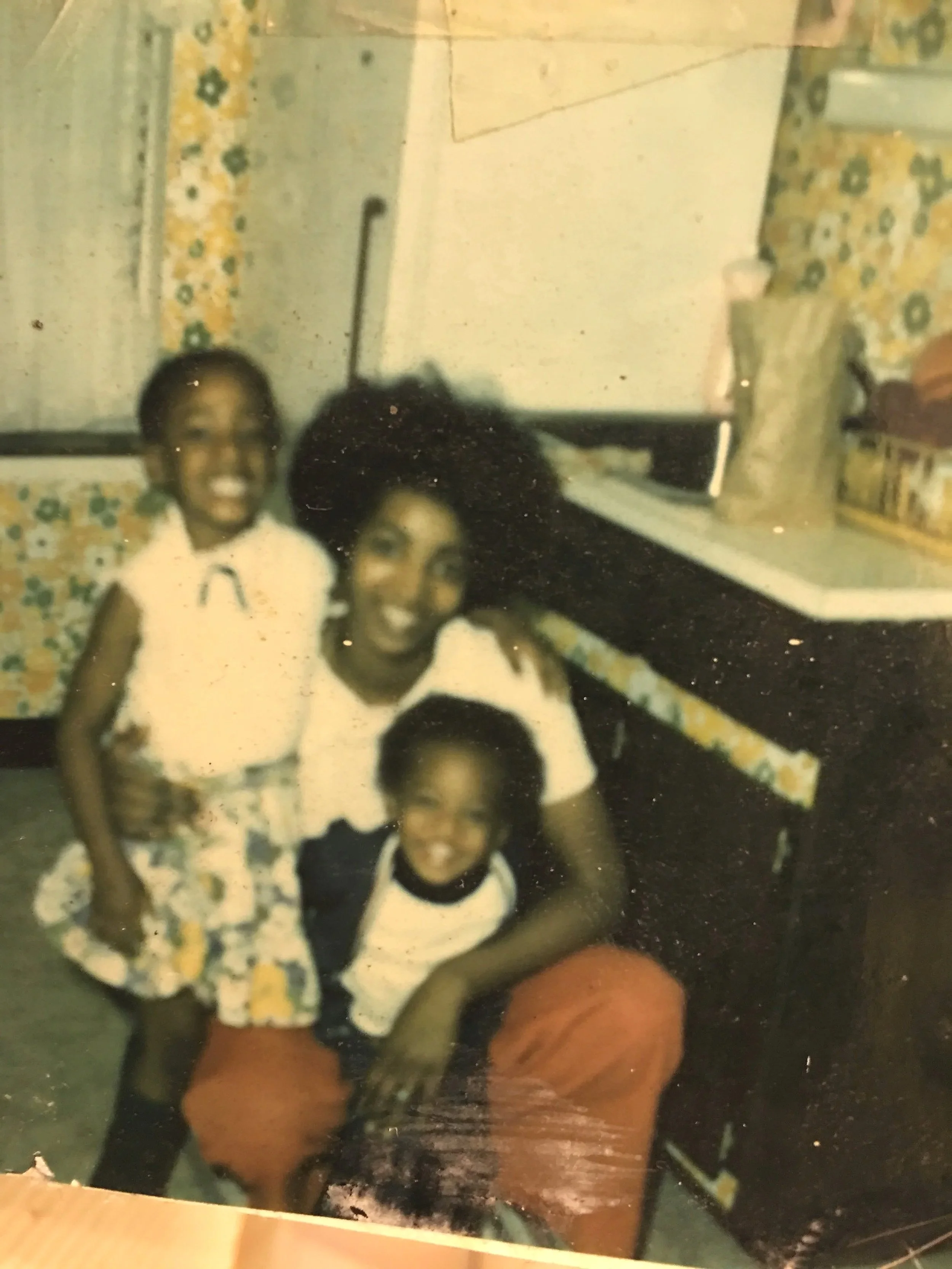 A woman with curly hair sitting on a chair, T-shirt, and orange pants, smiling. Two young girls with similar hairstyles and floral skirts, also smiling, standing around her. The background has floral-patterned curtains, a counter with various items, and a whiteboard.