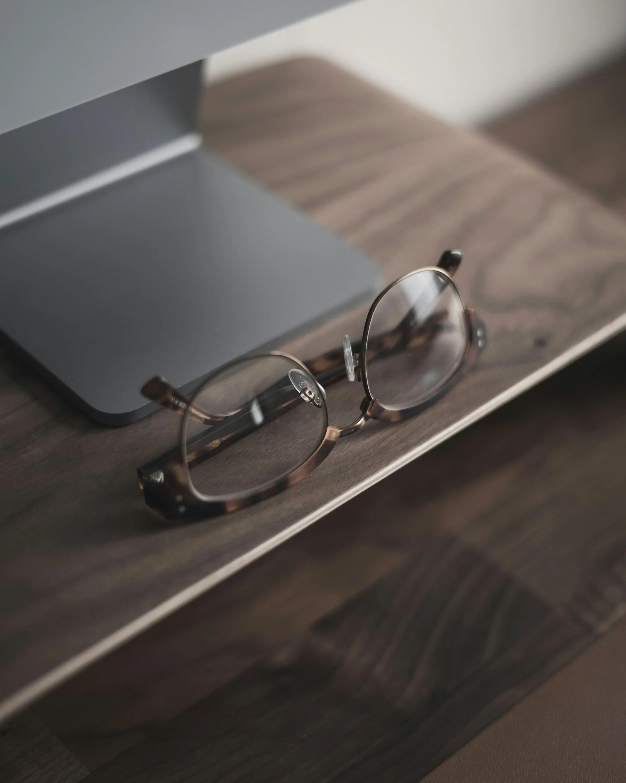 Wooden executive desk with eyeglasses with a metallic frame resting on its surface, partially under a gray electronic device.