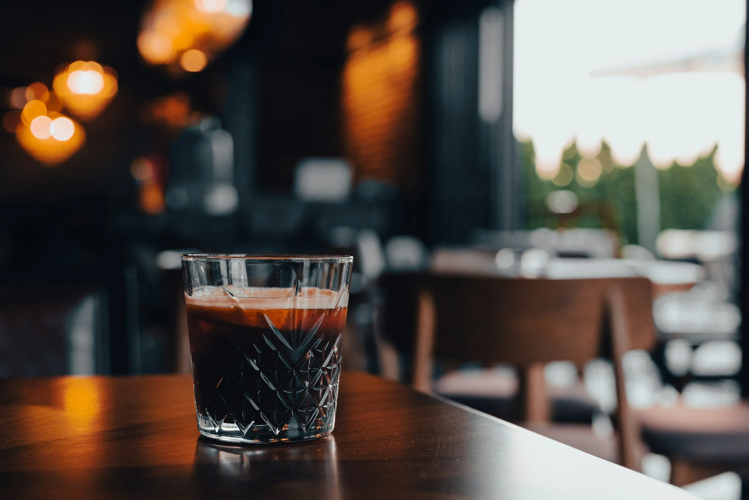 A glass of dark espresso coffee on a wooden table in a cozy, dimly lit cafe with blurred chairs and windows in the background.