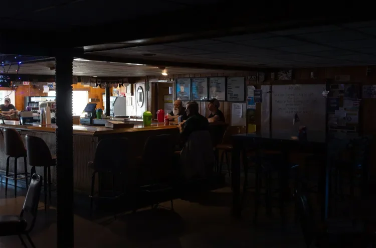 A dimly lit bar or pub with customers sitting at the counter and a few people in the background, posters and menus on the wall.