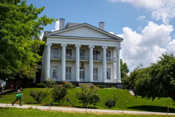 Large white historic house with tall columns, balcony, and manicured lawn.