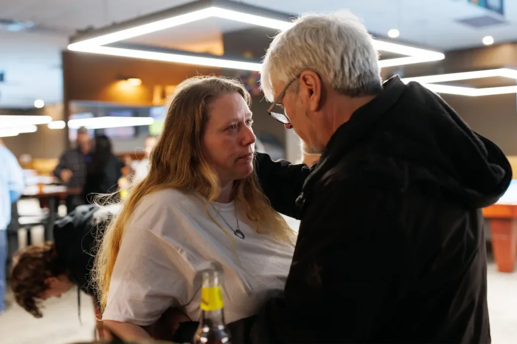 A woman and an older man share an emotional moment indoors, with the man touching the woman's face and both looking at each other, surrounded by warm indoor lighting.