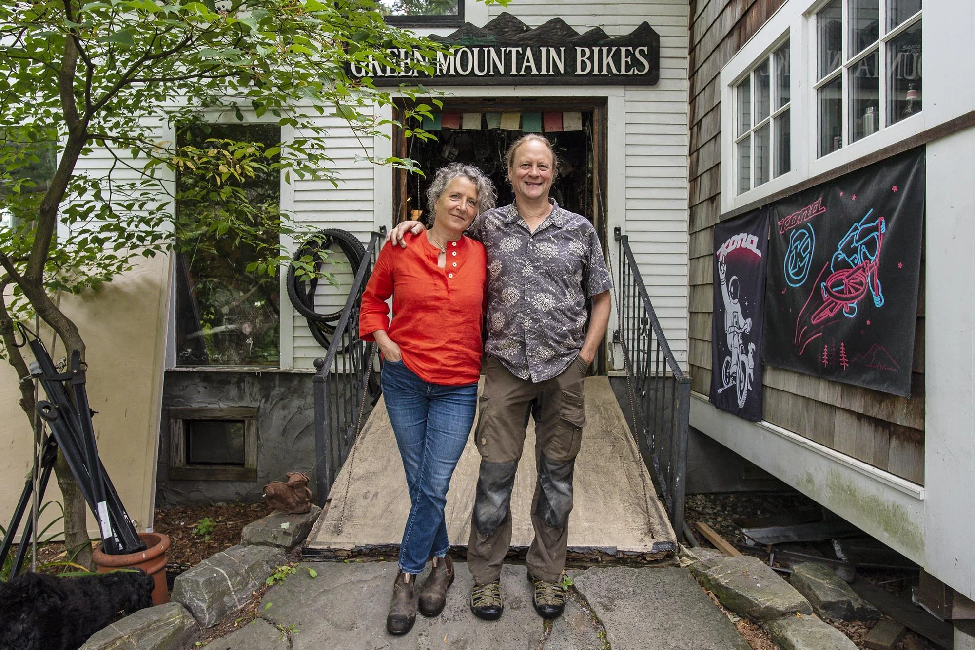A man and woman standing together on a wooden ramp outside a shop named GREEN MOUNTAIN BIKES, with mountain bike graphics on the wall to the right. The woman is wearing a red shirt and jeans, and the man is wearing a patterned shirt and pants. They are smiling and have their arms around each other, with greenery and trees surrounding the scene.