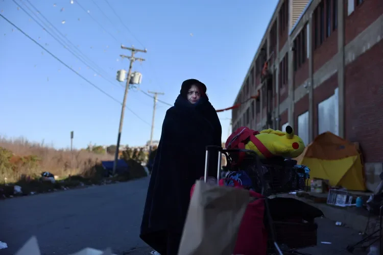 Person dressed in black clothing standing on a sidewalk next to a shopping cart filled with various items, including a plush toy, with a row of brick buildings and utility poles in the background.