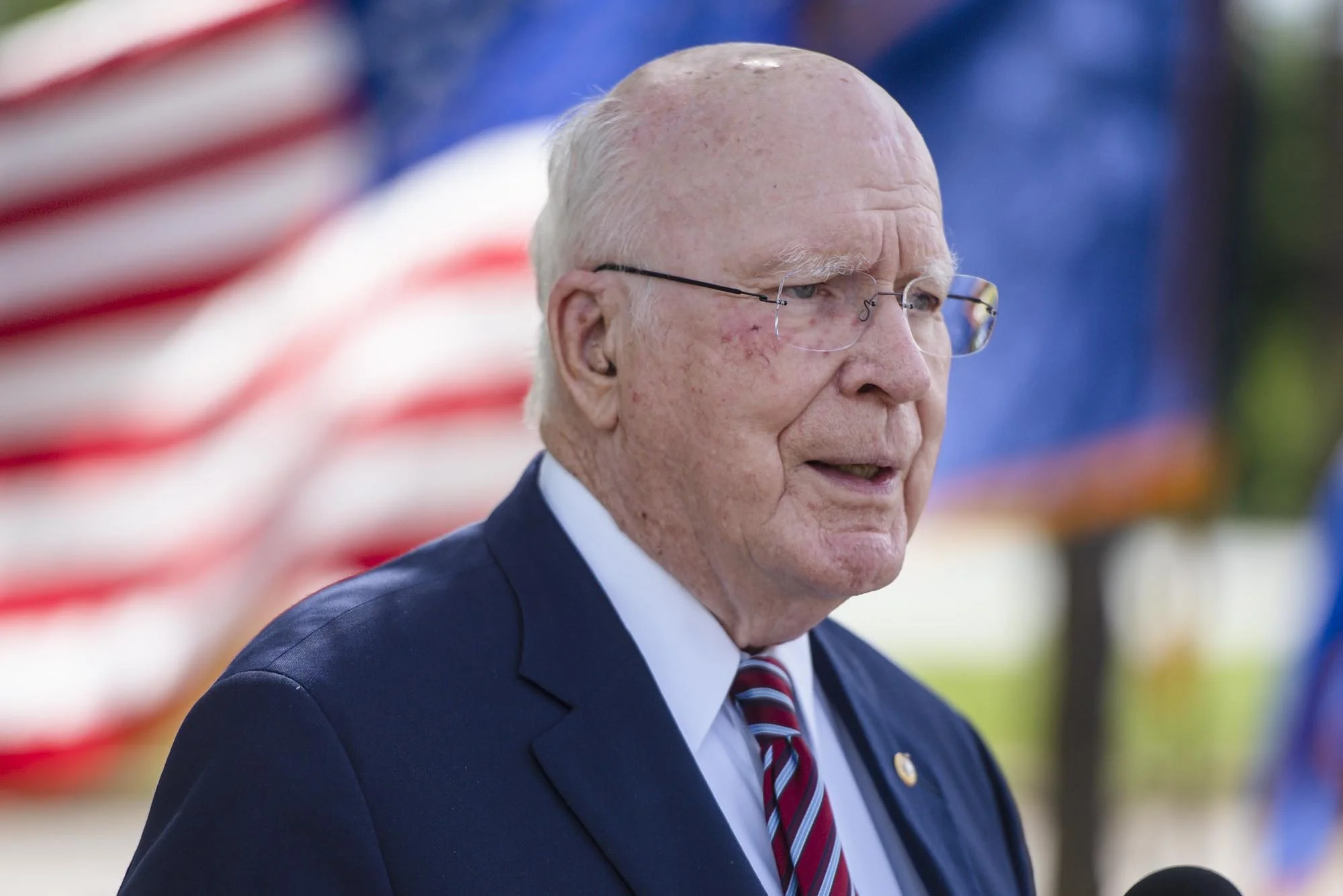 An elderly man with glasses and white hair, wearing a navy suit, white shirt, and red striped tie, speaking outdoors with an American flag in the background.