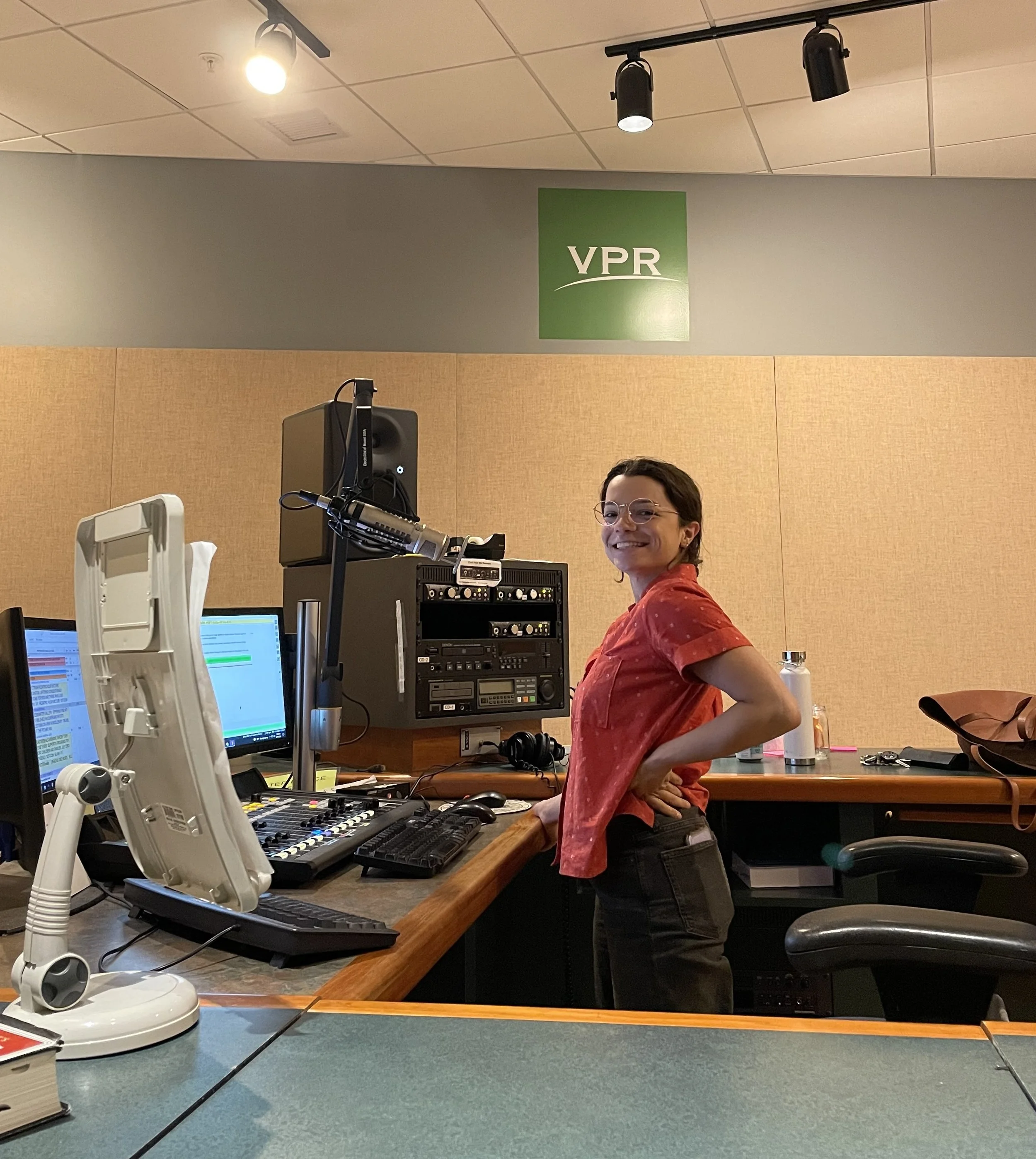 A woman working in a radio studio, standing at a console with multiple computer screens, microphones, and recording equipment, with a green VPR sign on the wall behind her.