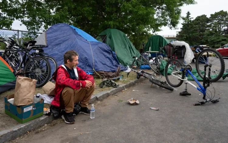 Man sitting on the sidewalk near tents and bicycles in a homeless encampment.