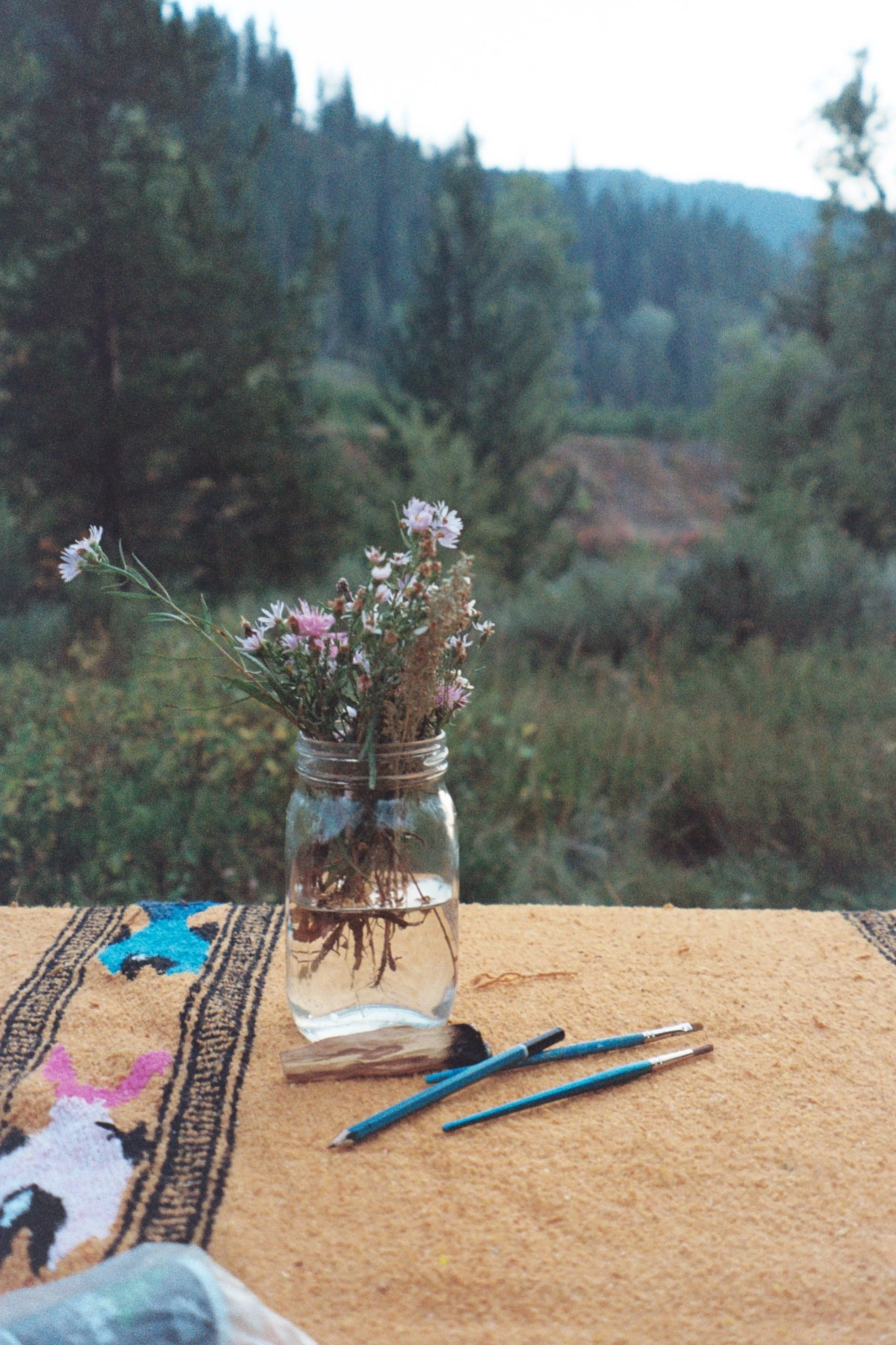 A jar with water containing a bouquet of wildflowers on a yellow textile with patterns, set outdoors with a background of trees and mountains.