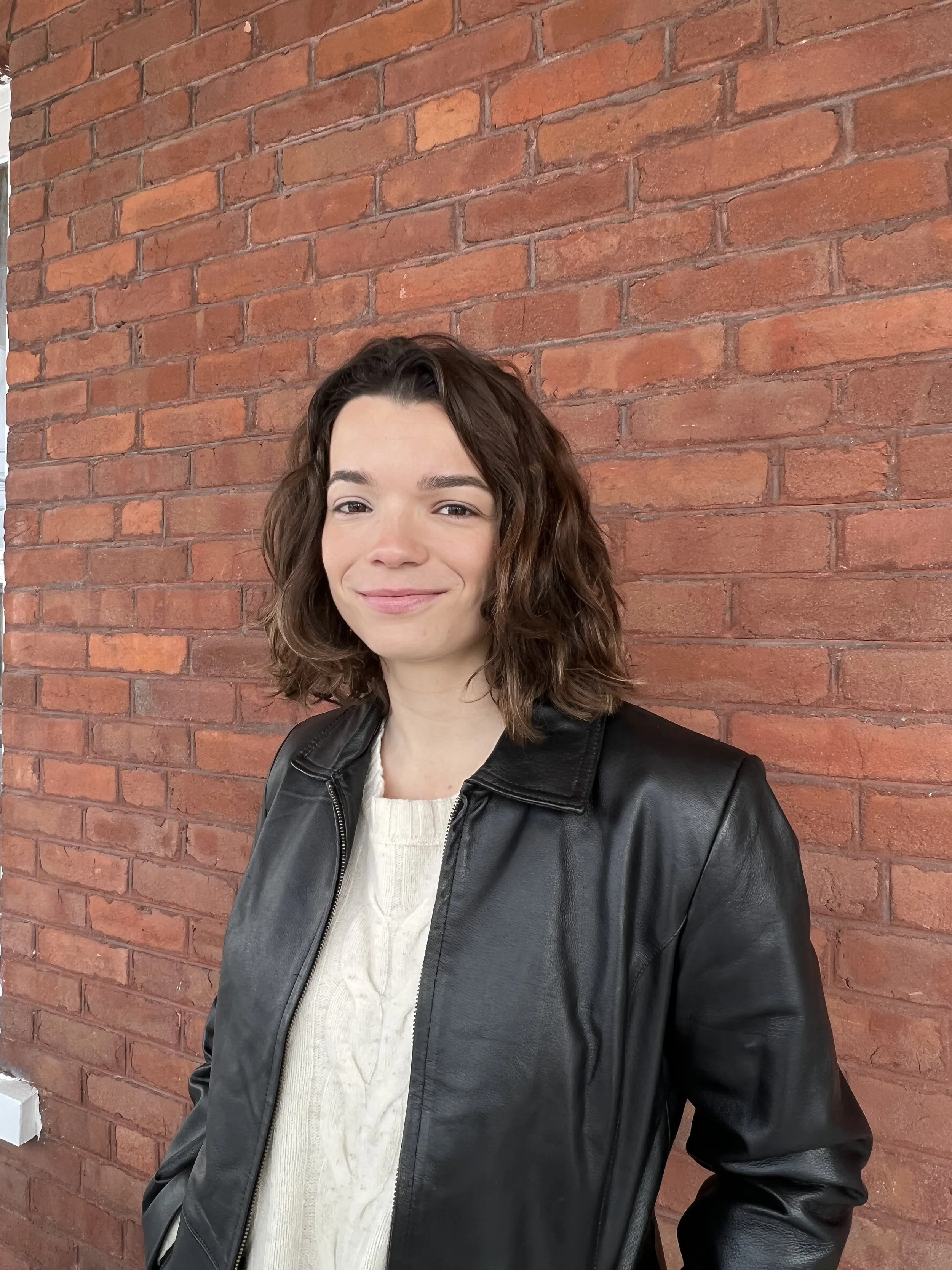 A woman with shoulder-length wavy brown hair, wearing a black leather jacket over a cream cable knit sweater, standing in front of a red brick wall.