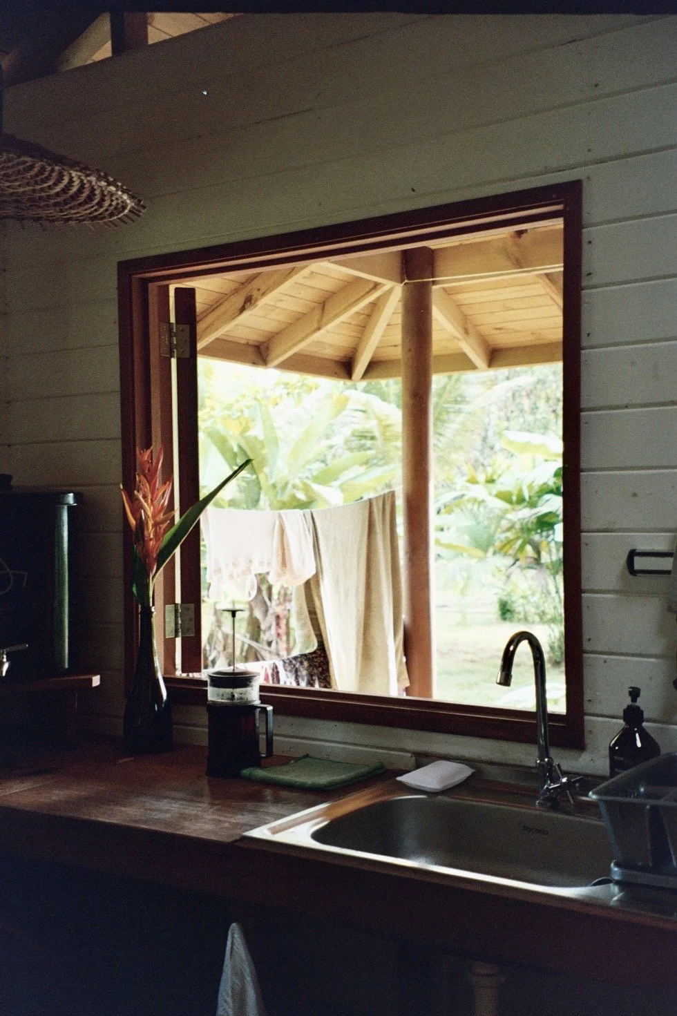 View from inside a kitchen looking out through a large open window to a porch with a thatched roof and leafy outdoor scenery.