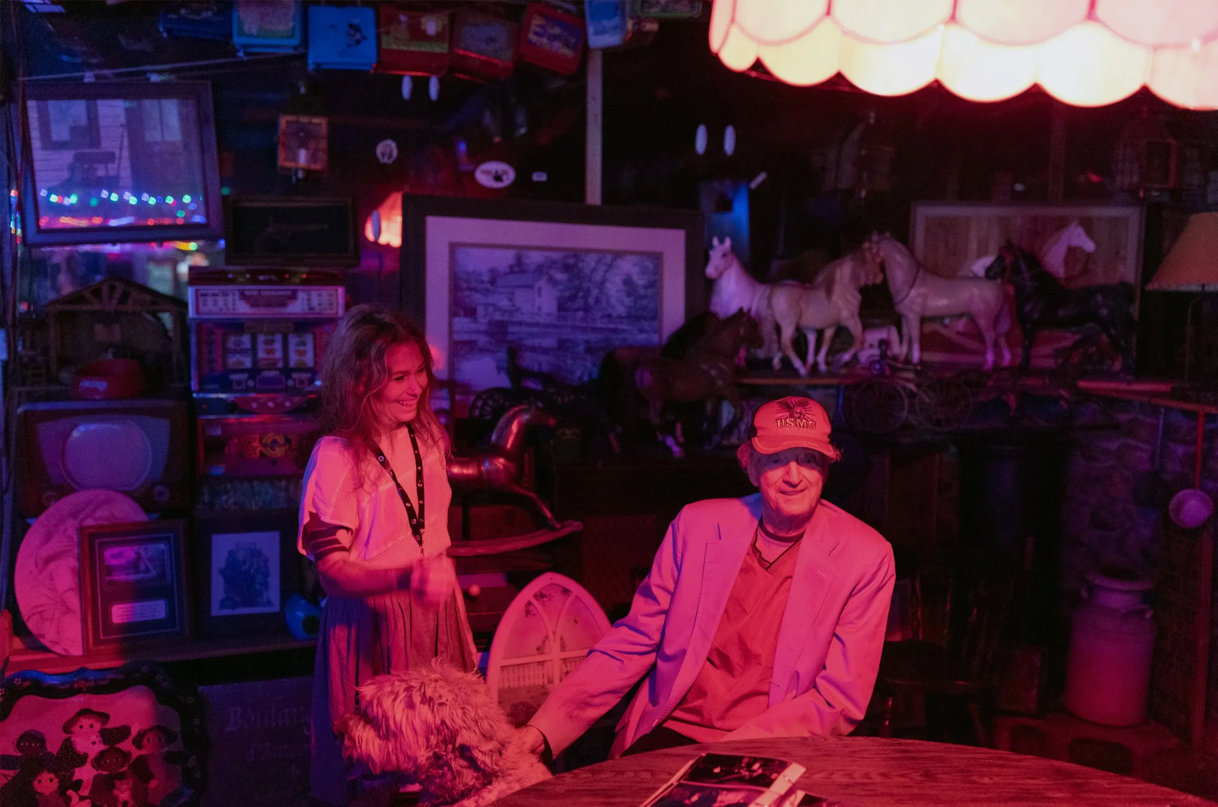 A woman and an older man in a room with vintage toys and decor, including model horses and framed pictures, illuminated by pink and purple lighting, with the woman smiling and the man looking at the camera.