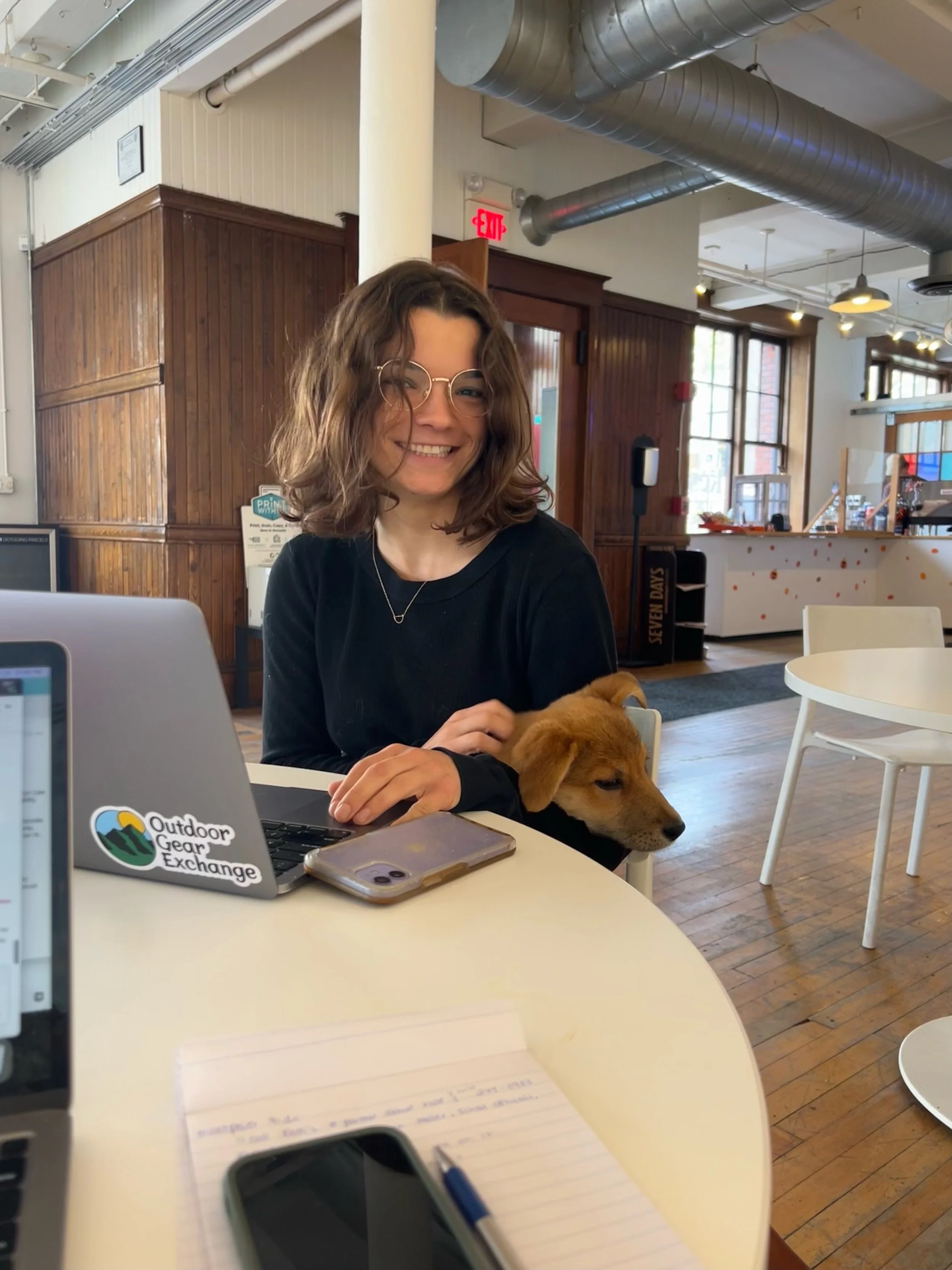 Smiling woman with curly brown hair and glasses sitting at a table in a coffee shop with a small brown puppy, surrounded by laptops, phones, and notes.