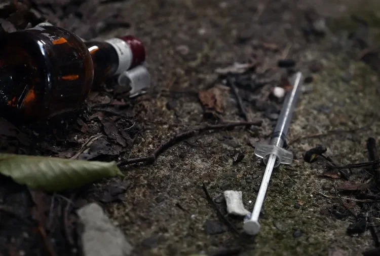 A used medical syringe lying on the ground next to a broken medicine bottle and other debris on dirt and leaves.