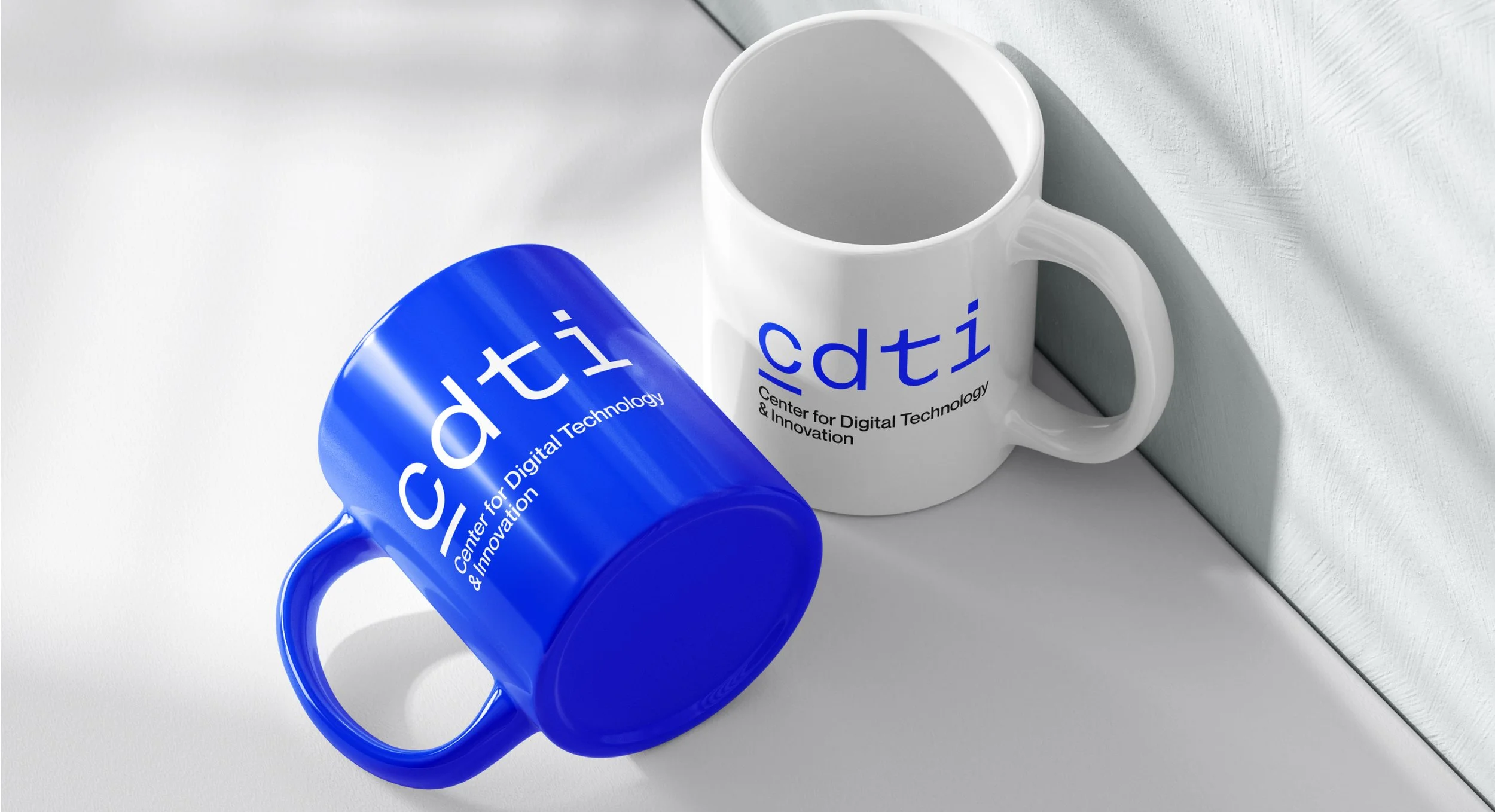 Two ceramic mugs, one blue and one white, featuring the logo of the Center for Digital Technology & Innovation, placed on a surface with a white textured wall behind them.