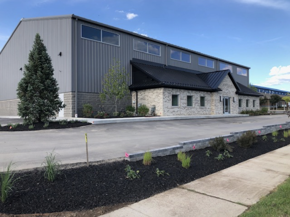 A modern commercial building with a combination of metal siding and stone facade, black roof, and several small windows. The parking area is freshly paved with new landscaping and small plants along the edge, under a partly cloudy blue sky.