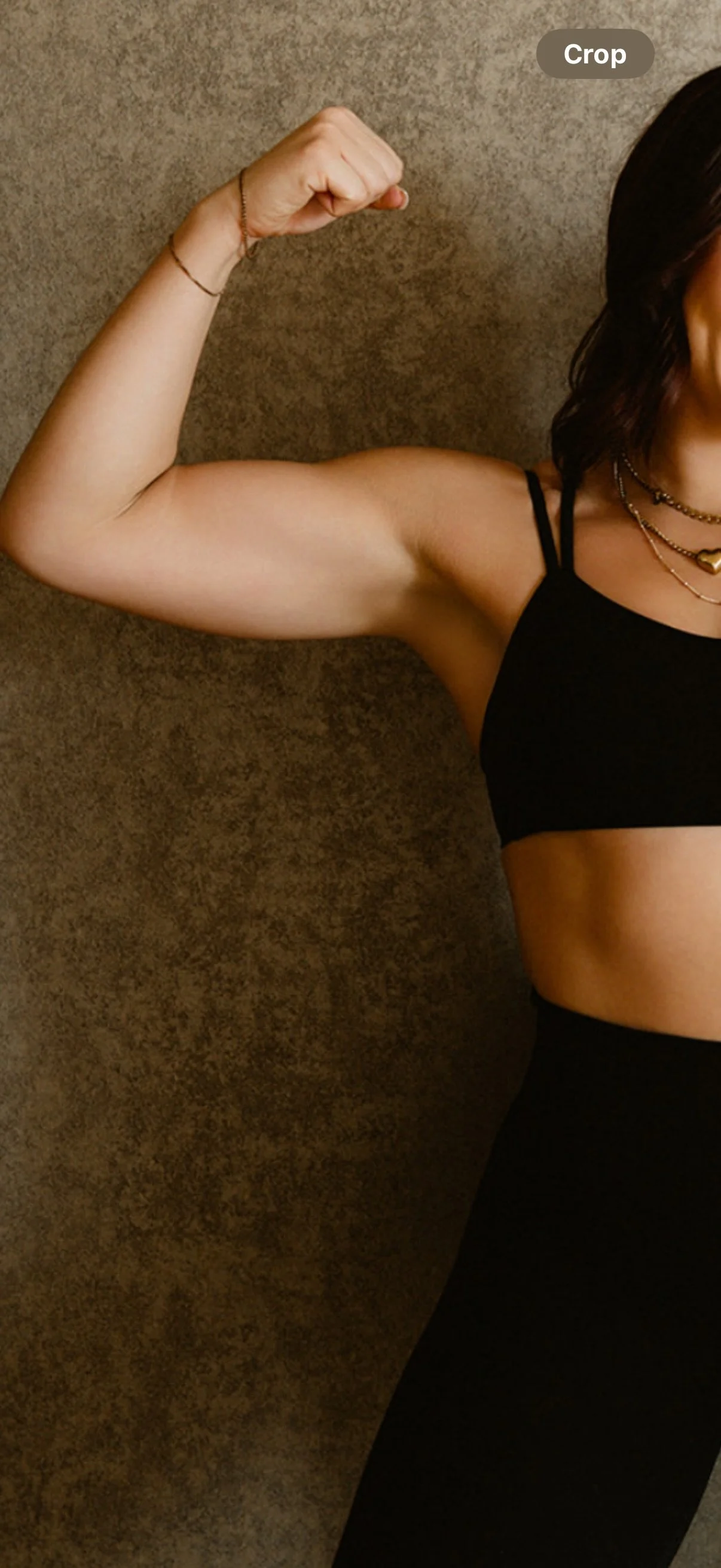 A woman flexing her bicep muscle, wearing a black sports bra and black leggings, with layered necklaces, standing against a textured brown wall.