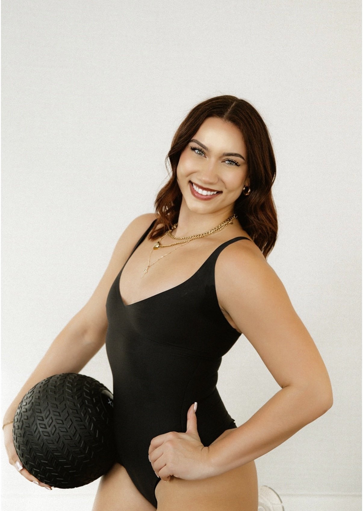 A young woman with shoulder-length brown hair, wearing a black workout top, holding a black textured medicine ball, smiling at the camera, standing against a plain white wall.