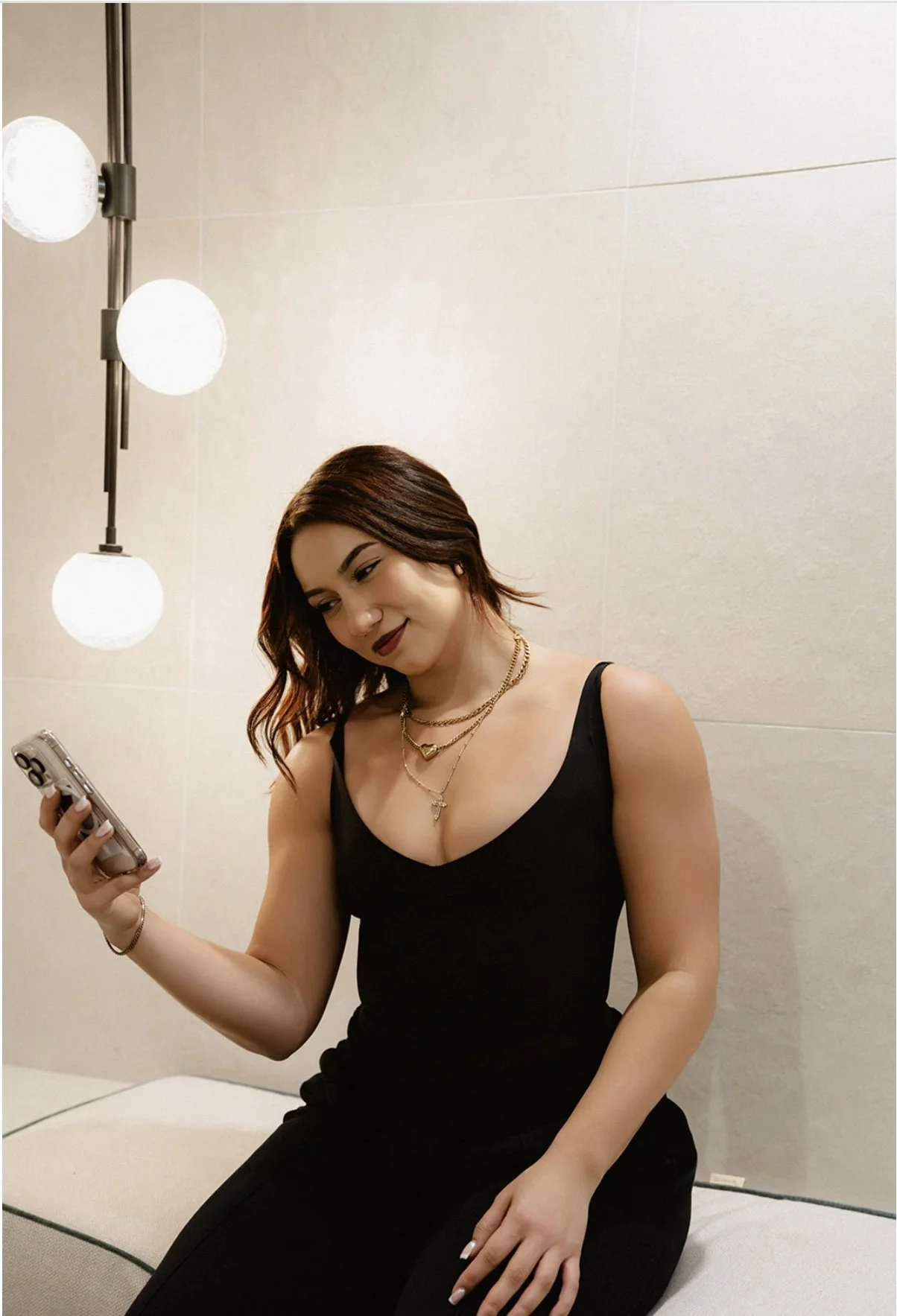 A woman with shoulder-length brown hair, wearing a black sleeveless top and multiple gold necklaces, sitting on a beige cushioned bench looking at her smartphone in a modern interior with beige tiles and a wall-mounted light fixture.