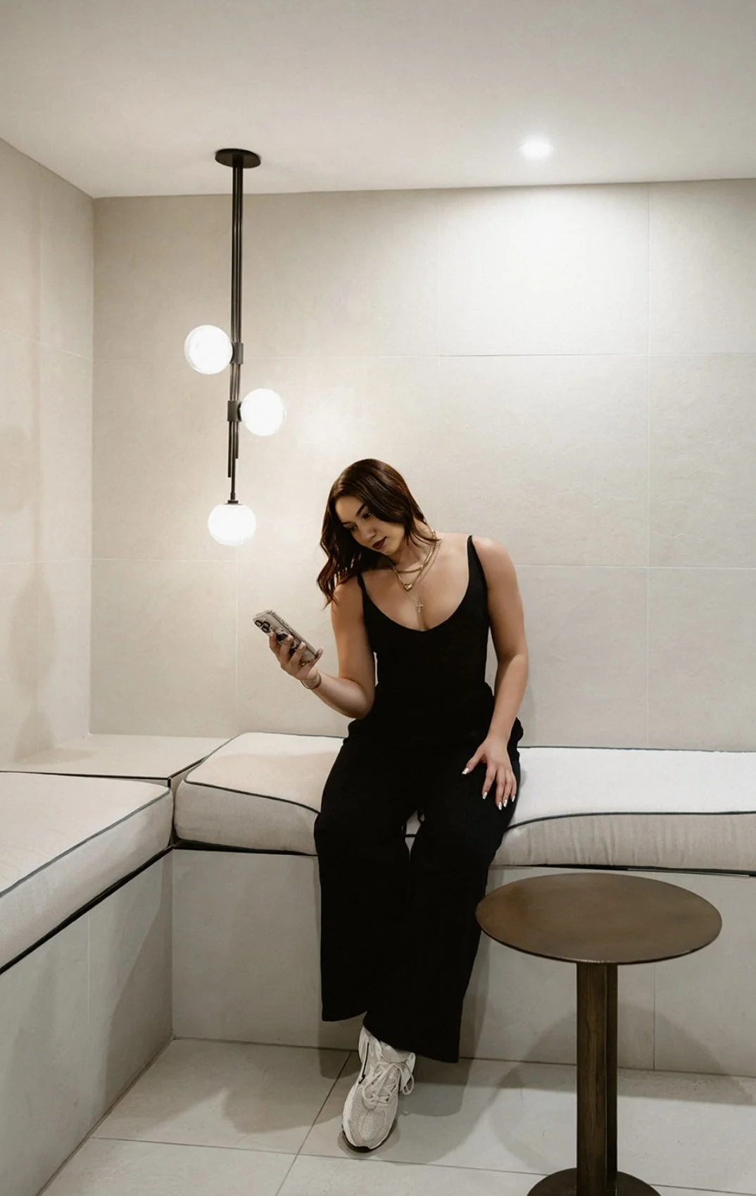 Woman in black dress sitting on white cushioned bench in a modern, minimalistic room, looking at her phone.