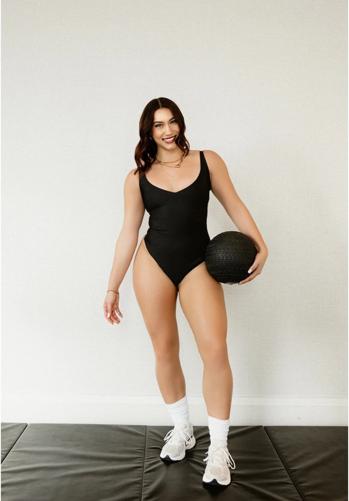 Woman smiling in black leotard holding a black medicine ball, wearing white athletic shoes and socks, standing on a black mat against a plain white background.