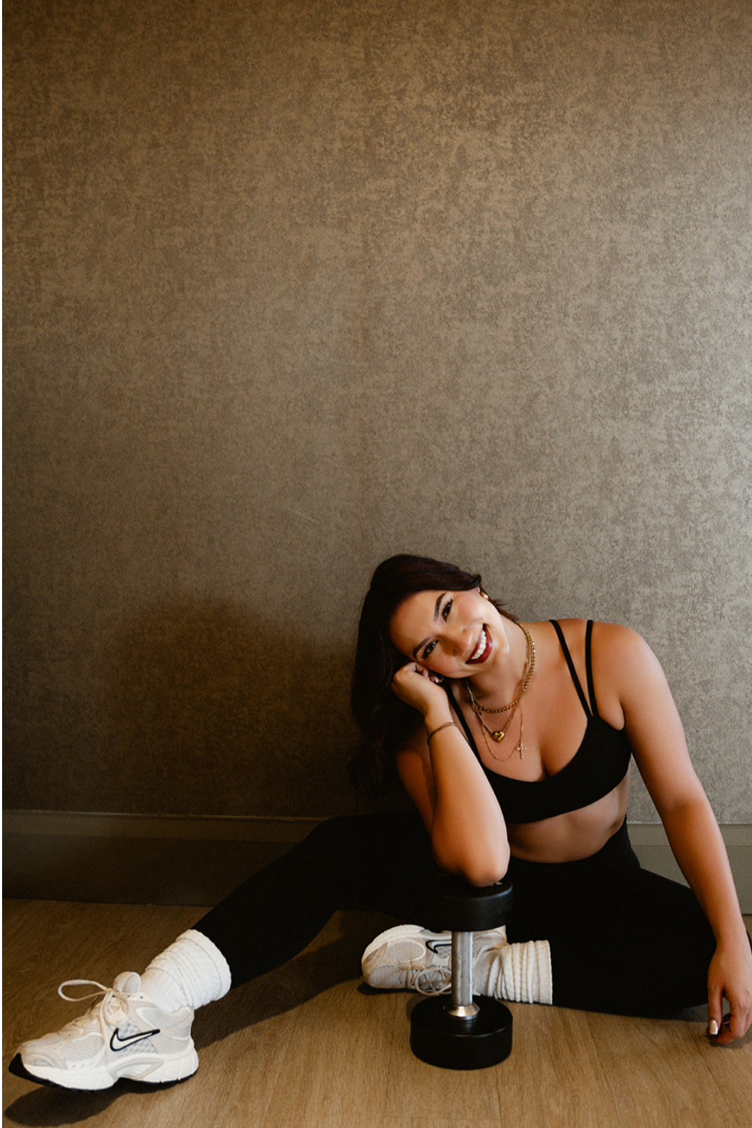 A woman with dark hair sitting on the floor, smiling, with her head resting on her hand, next to a dumbbell and wearing workout clothes.