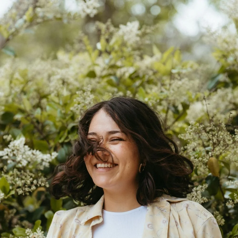 A woman smiling with curly dark hair outdoors in front of green leafy bushes with white flowers.