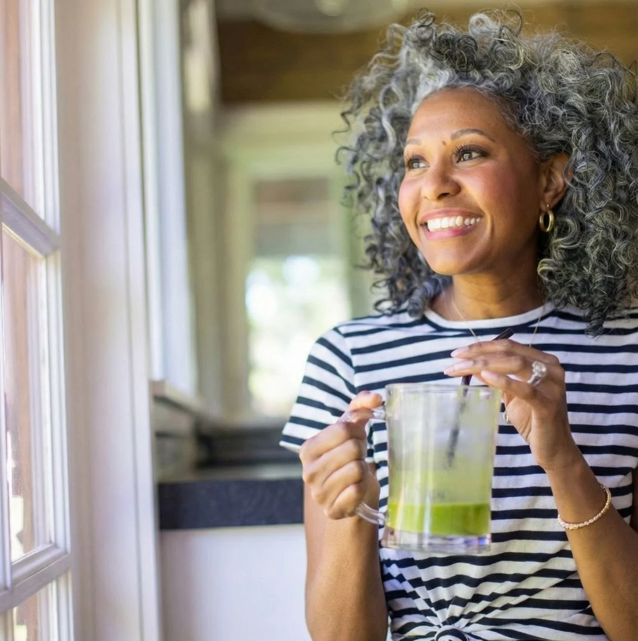 A woman with curly gray hair smiling and looking out a window while holding a glass of green juice.