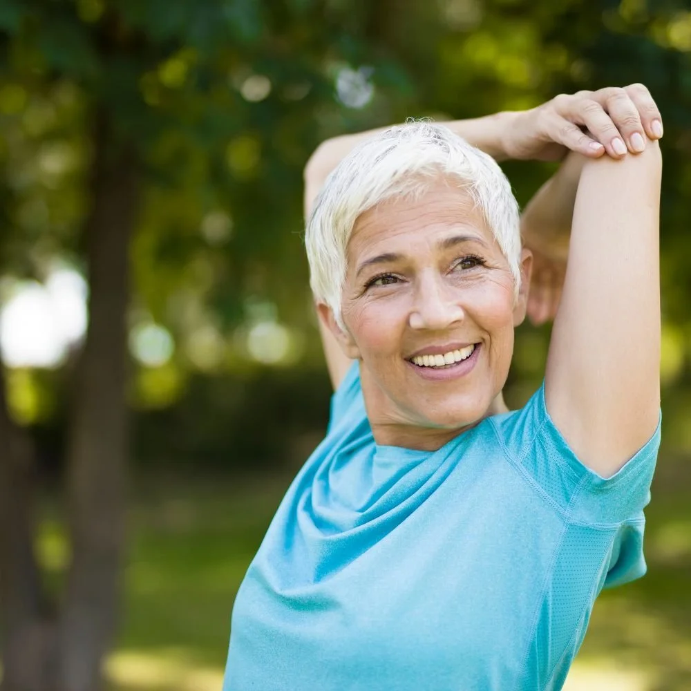An elderly woman stretching outdoors in a park, smiling and looking happy.