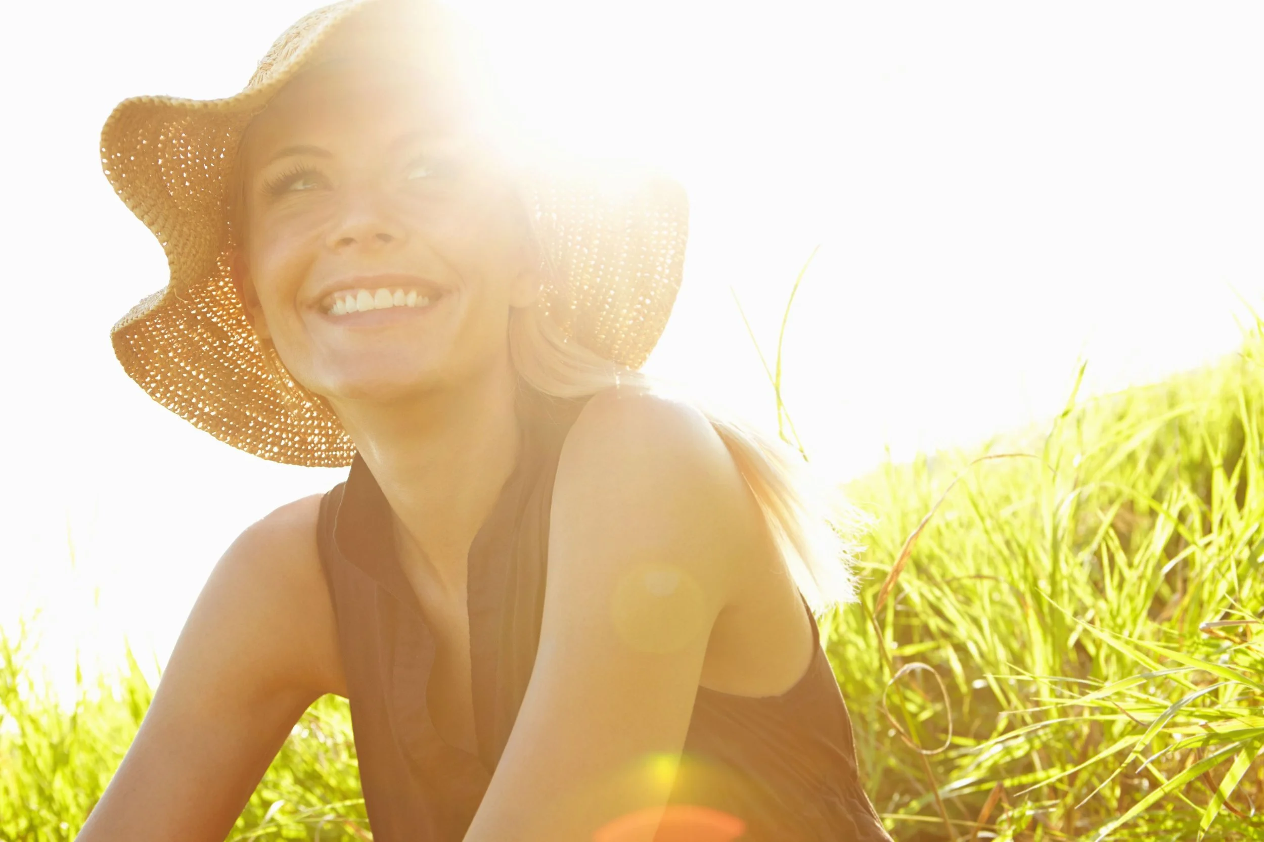 A woman smiling and wearing a wide-brimmed straw hat outdoors in a sunlit field of tall grass.
