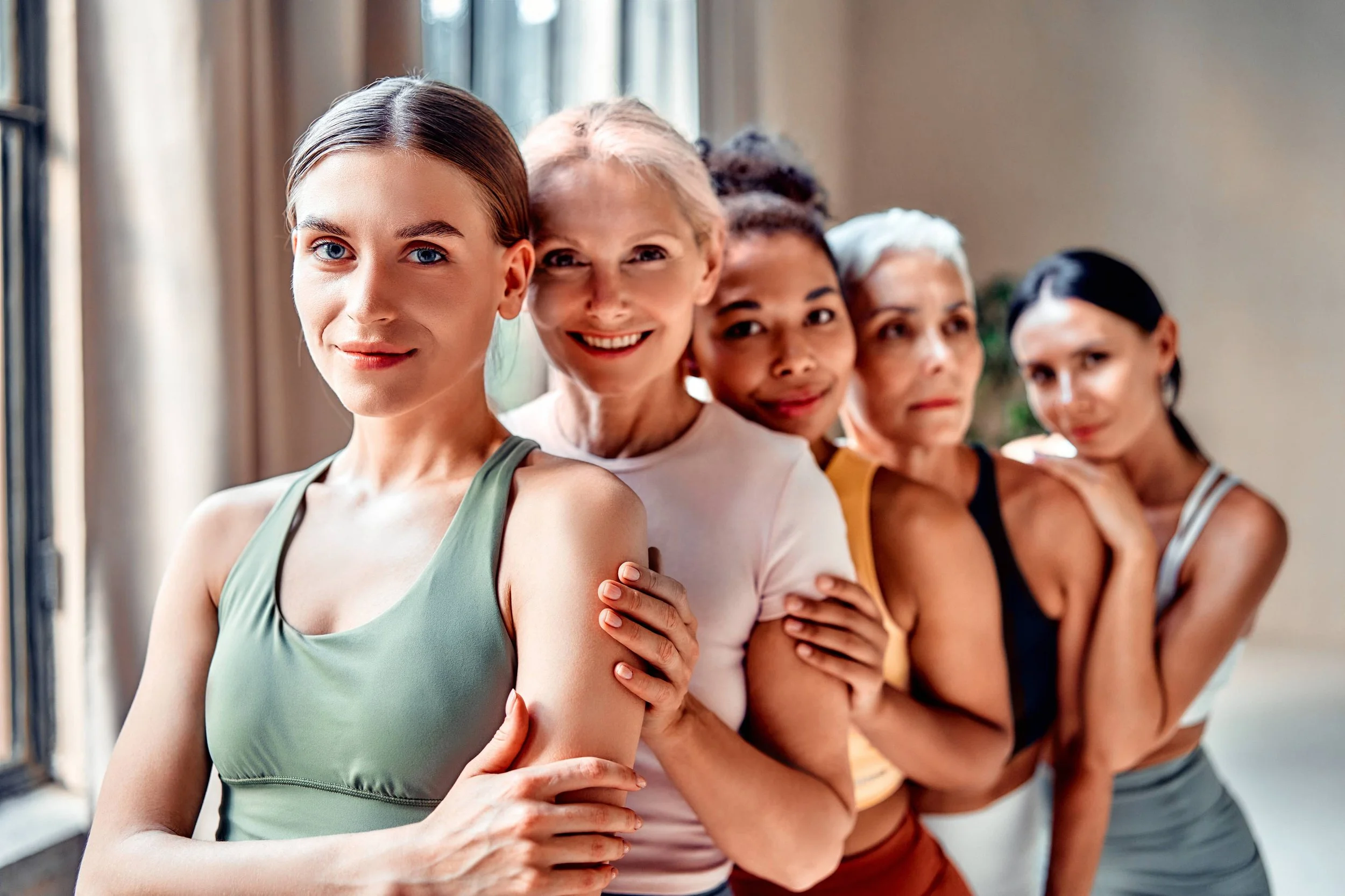 Five women of diverse ages standing in a line indoors near a window, smiling and hugging each other, dressed in workout or casual clothes.