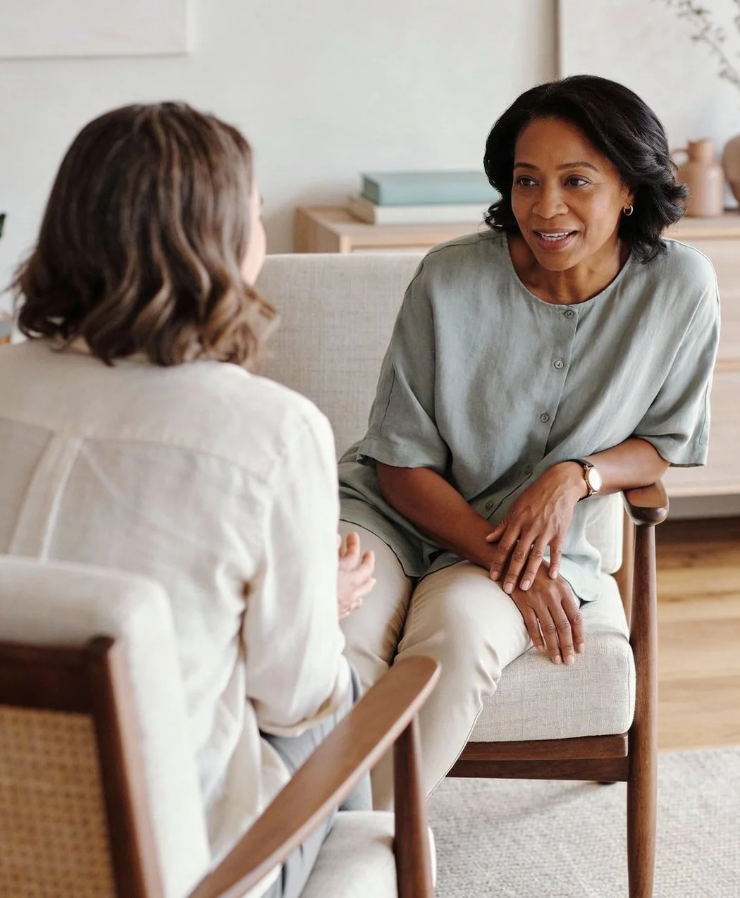 Two women having a conversation in a cozy room with a beige sofa and wooden furniture.